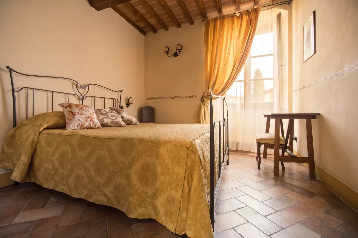Camera Porta Senese: letto matrimoniale in ferro battuto, copriletto damascato, tende ocra e soffitto a travi
